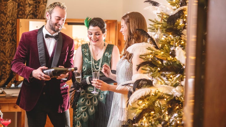 A festive scene with three people in vintage attire. A man in a velvet jacket pours champagne for two women near a decorated, illuminated tree.